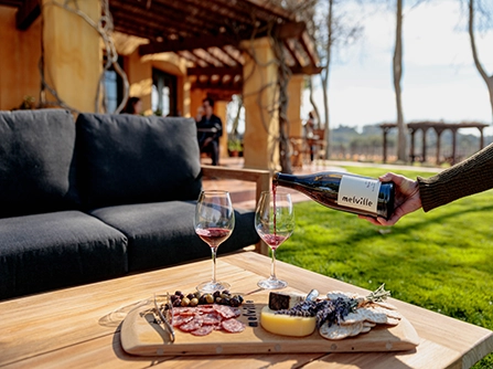 Hand pouring red wine beside a small charcuterie tray of meats and cheeses at a Santa Barbara winery with outdoor seating.